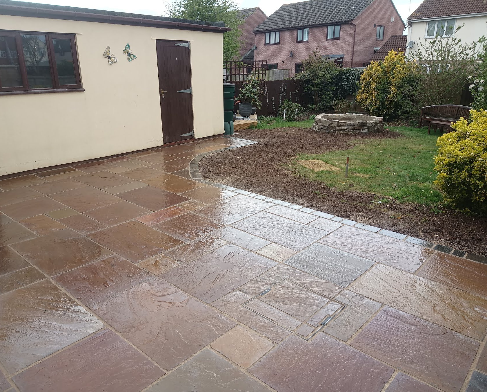 A new sandstone patio beside a rendered outbuilding, with a fresh soil bed and lawn behind