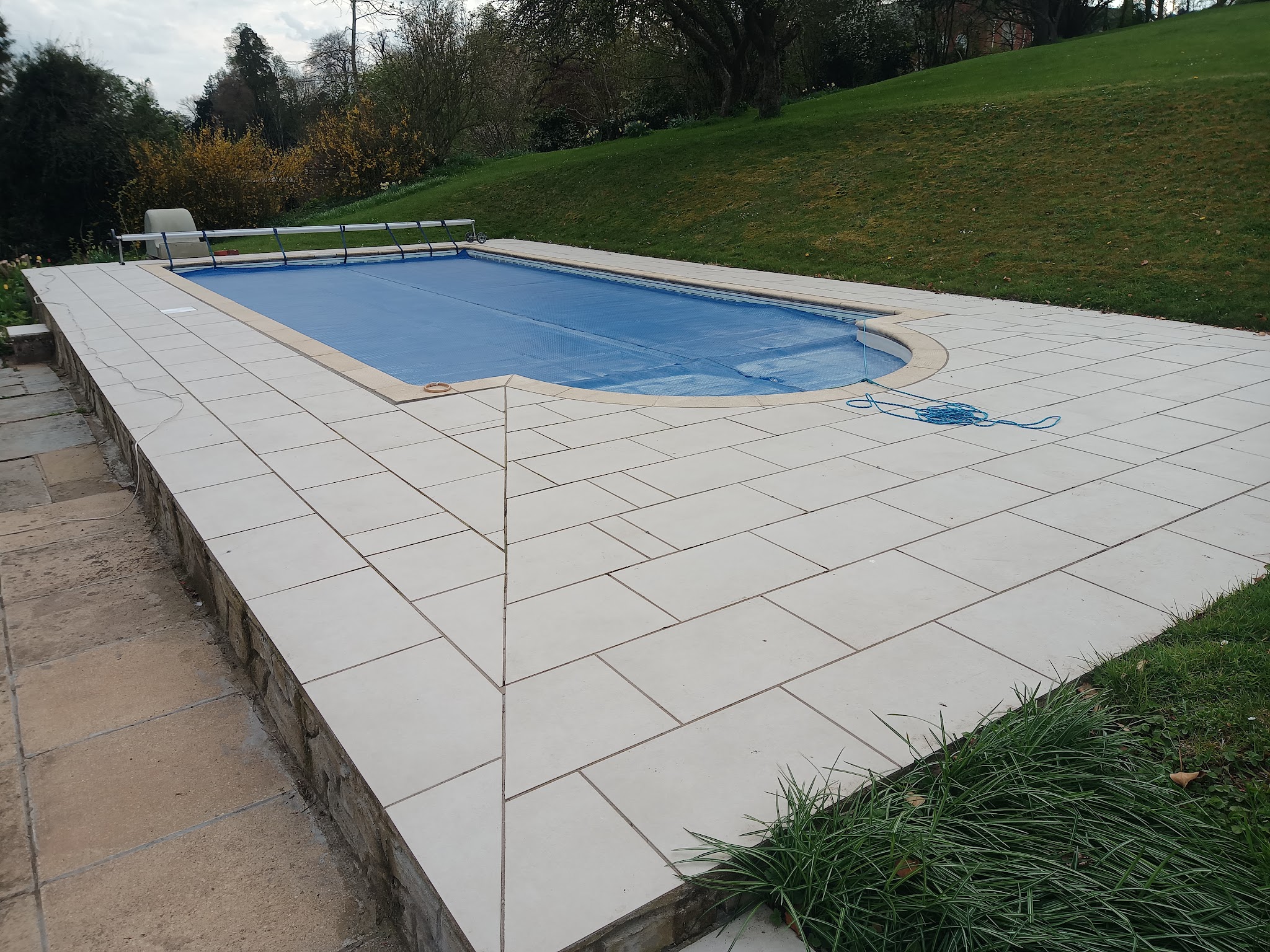Pale paving slabs laid around a swimming pool on a sloping lawn
